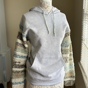 May include: A grey hooded sweatshirt with crocheted sleeves in shades of brown, blue and white.