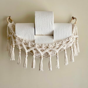 May include: A white crocheted hammock-style toilet paper holder with wooden beads and tassels. Three rolls of toilet paper are in the holder.