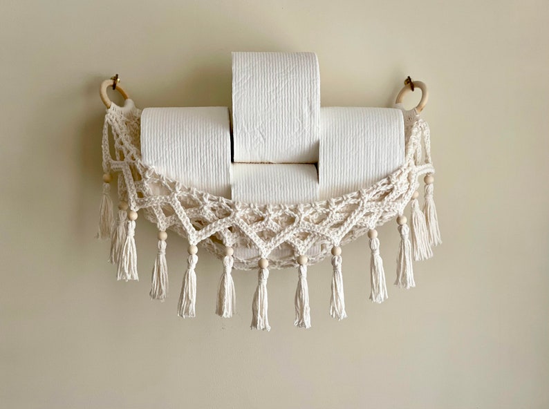 May include: A white crocheted hammock-style toilet paper holder with wooden beads and tassels. Three rolls of toilet paper are in the holder.