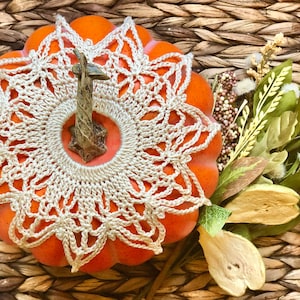 May include: A small orange pumpkin with a beige crocheted doily on top. The pumpkin is sitting on a woven wicker surface.