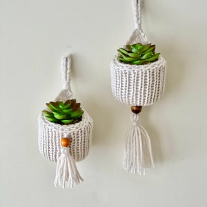 May include: Two white macrame hanging planters with wooden beads and tassels. Each planter holds a small green succulent plant.