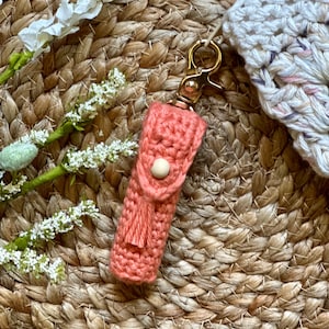 May include: A coral crocheted keychain with a tassel and a wooden button. The keychain is attached to a gold metal clasp.