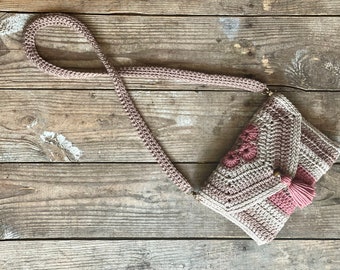 May include: A handmade crochet shoulder bag in shades of taupe and dusty rose. The bag features a flap closure, decorative floral accents, and a tassel. The strap is also crocheted. The bag is displayed on a rustic wooden surface.