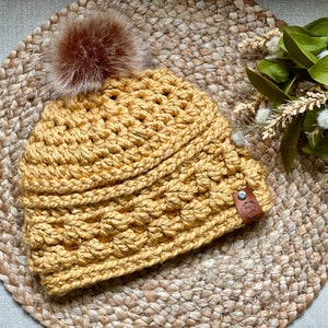 May include: A mustard yellow crocheted beanie with a brown faux fur pom pom on top. The beanie has a brown leather tag with the text "The Cozy Co."