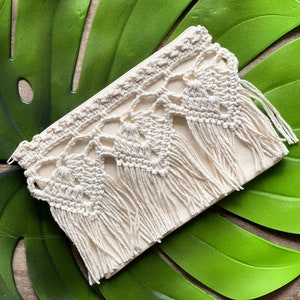 May include: A white crocheted pouch with a fringe border. The pouch is lying on a green leaf.