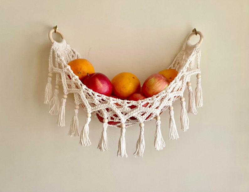 May include: A white crocheted hammock-style fruit basket with wooden beads and tassels hanging from a wall. The basket is filled with red apples and orange oranges.