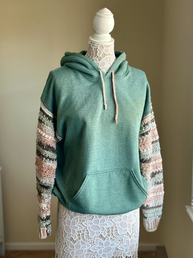 May include: A green hooded sweatshirt with crocheted sleeves in shades of brown, pink, and white.