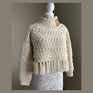 May include: A cream-coloured, chunky knit sweater with a high collar and fringe detail. The sweater has two brown buttons on the shoulder and a textured pattern. The sleeves are long, and the hem has a fringe.