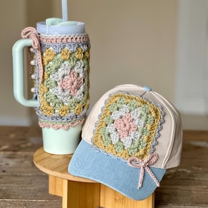 May include: A light green water bottle with a straw and a matching crochet cosy, next to a baseball cap. The cosy and cap feature a square crochet design in pastel colours. The cap has a blue brim and a pink bow.