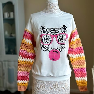 May include: White sweatshirt featuring a tiger wearing pink sunglasses and blowing a pink bubble. The sleeves are crocheted with stripes of pink, orange, yellow, and white.