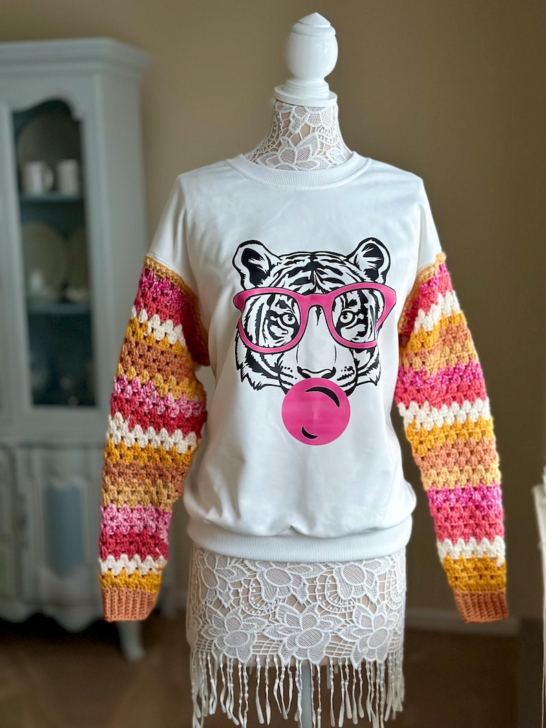 May include: White sweatshirt featuring a tiger wearing pink sunglasses and blowing a pink bubble. The sleeves are crocheted with stripes of pink, orange, yellow, and white.