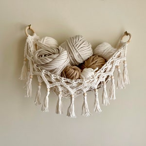 May include: A white crocheted hammock with wooden beads and tassels hangs on a wall. The hammock is filled with balls of yarn in various shades of white and beige.