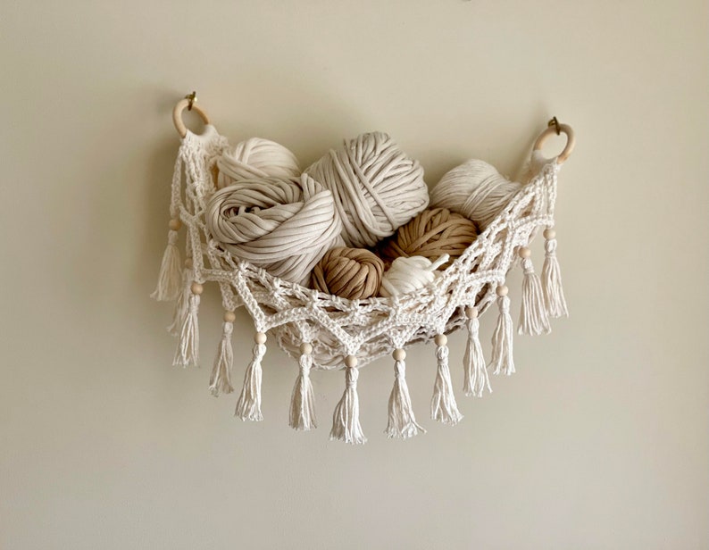 May include: A white crocheted hammock with wooden beads and tassels hangs on a wall. The hammock is filled with balls of yarn in various shades of white and beige.