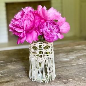 May include: A bouquet of pink peonies in a glass jar with a crocheted cover and fringe.