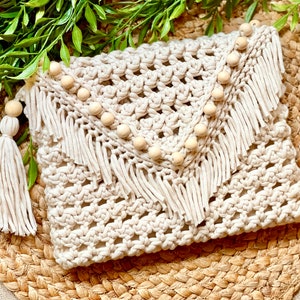 May include: A crocheted clutch bag with a fringe trim and wooden beads. The bag is beige and has a woven texture.