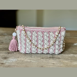 May include: A handmade crochet bag in a rectangular shape, featuring alternating vertical stripes of light pink and off-white. It has a gold chain strap and a pink tassel with a small white pom-pom. The bag is on a wooden surface.