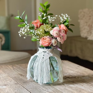 May include: A white crocheted vase cover with fringe, holding a bouquet of pink and white flowers. The vase is clear glass and the flowers include baby's breath, pink roses, and pink carnations.