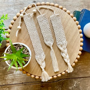 Puede incluir: Tres marcapáginas de macramé hechos a mano con borlas de color crema natural. Los marcapáginas se muestran en una bandeja de madera con cuentas decorativas.