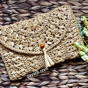 May include: A beige raffia clutch bag with a woven floral pattern. It has a flap closure with a wooden bead and tassel. The website address daysck.com is visible. The bag is set against a textured, woven background.