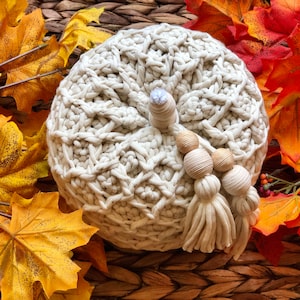May include: A cream-colored crocheted pumpkin with a tassel and wooden beads. The pumpkin is surrounded by fall leaves in shades of orange and yellow.