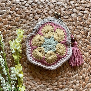 Puede incluir: Un monedero de ganchillo con un diseño floral en tonos beige, rosa y verde. Tiene un cierre metálico y una borla. El fondo es una textura tejida.