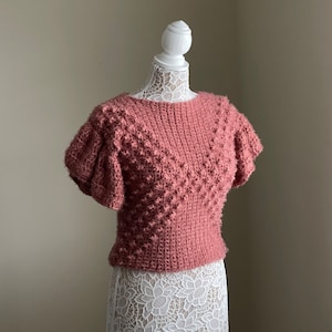 May include: A dusty rose crocheted top with a textured, bobble stitch pattern. The top has short, puffed sleeves and a round neckline.