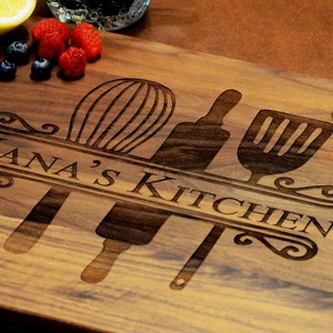May include: A wooden cutting board with the text "Nana's Kitchen" engraved on it. The board is decorated with three kitchen utensils: a whisk, a rolling pin, and a spatula. The board is surrounded by fruit, including kiwi slices and orange slices.