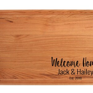 K&ouml;nnte beinhalten: Ein Schneidebrett aus Holz mit abgerundeter Kante und einer Rille um den Umfang. Das Brett ist mit dem Text "Welcome Home Jack & Hailey Est. 2018" graviert.