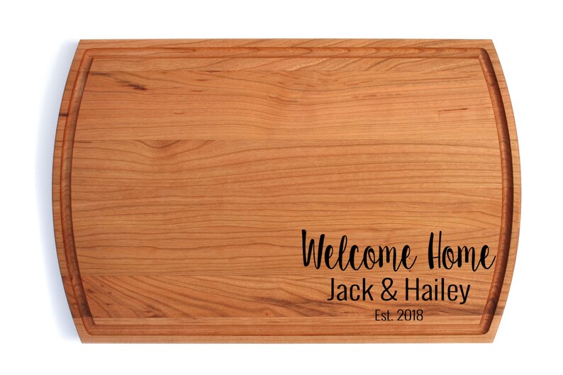 K&ouml;nnte beinhalten: Ein Schneidebrett aus Holz mit abgerundeter Kante und einer Rille um den Umfang. Das Brett ist mit dem Text "Welcome Home Jack & Hailey Est. 2018" graviert.