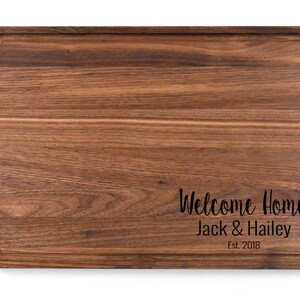 K&ouml;nnte beinhalten: Ein gro&szlig;es, rechteckiges, h&ouml;lzernes Schneidebrett mit abgerundeten Kanten. Das Brett ist aus Walnussholz gefertigt und hat eine nat&uuml;rliche Oberfl&auml;che. Das Brett ist mit dem Text "Welcome Home Jack & Hailey Est. 2018" graviert.