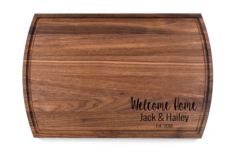 K&ouml;nnte beinhalten: Ein gro&szlig;es, rechteckiges, h&ouml;lzernes Schneidebrett mit abgerundeten Kanten. Das Brett ist aus Walnussholz gefertigt und hat eine nat&uuml;rliche Oberfl&auml;che. Das Brett ist mit dem Text "Welcome Home Jack & Hailey Est. 2018" graviert.
