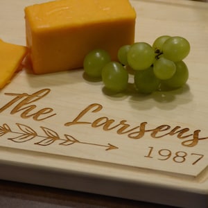 K&ouml;nnte beinhalten: Ein Holzbrett zum Schneiden mit einer personalisierten Inschrift, die "The Larsens 1987" lautet. Das Brett hat eine dekorative Pfeil- und Blattform. Auf dem Brett liegen ein St&uuml;ck Cheddar-K&auml;se und eine Traube gr&uuml;ner Trauben.