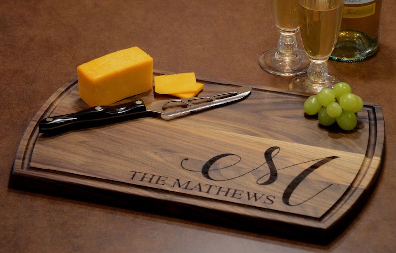 K&ouml;nnte beinhalten: Ein Schneidebrett aus Holz mit einer personalisierten Inschrift, die "Est. The Mathews" lautet. Das Brett ist braun und hat eine abgerundete Kante. Auf dem Brett liegen ein K&auml;semesser, ein St&uuml;ck K&auml;se und eine Traube gr&uuml;ner Trauben.