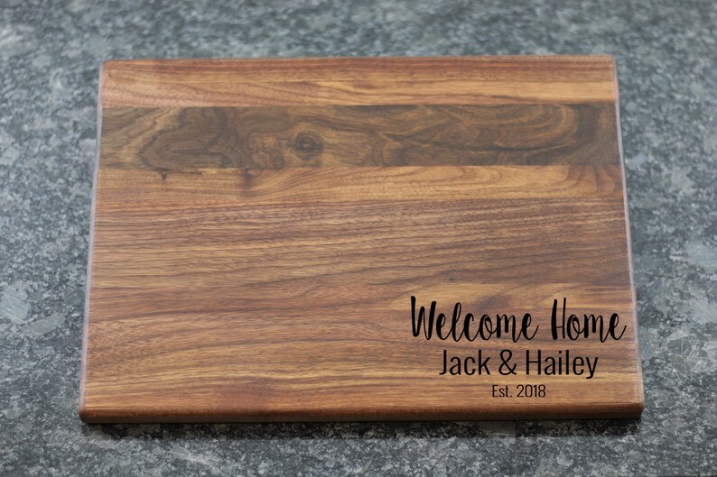 K&ouml;nnte beinhalten: Ein rechteckiges Schneidebrett aus Holz mit einer nat&uuml;rlichen Holzmaserung. Das Brett hat eine personalisierte Inschrift mit dem Text "Welcome Home Jack & Hailey Est. 2018".