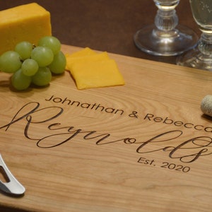 K&ouml;nnte beinhalten: Ein Schneidebrett aus Holz mit dem eingravierten Text "Johnathan & Rebecca Reynolds Est. 2020". Das Brett ist von K&auml;se, Trauben, einem Korkenzieher und einem Flaschen&ouml;ffner umgeben.