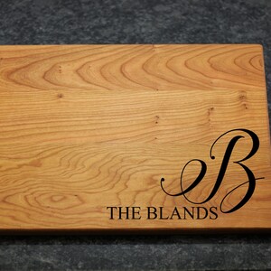 May include: A wooden cutting board with a natural wood finish. The board is engraved with the letter "B" in a cursive font and the text "THE BLANDS".