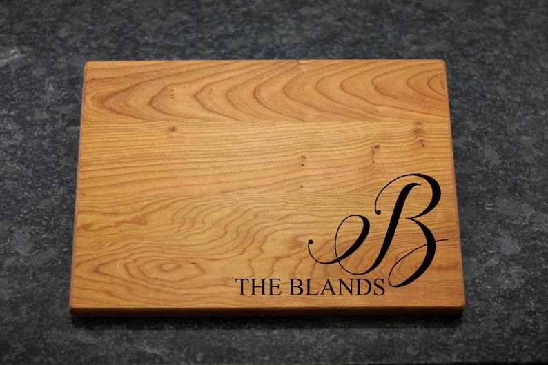 May include: A wooden cutting board with a natural wood finish. The board is engraved with the letter "B" in a cursive font and the text "THE BLANDS".