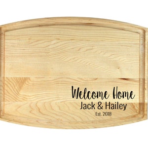 K&ouml;nnte beinhalten: Ein Schneidebrett aus Holz mit abgerundeten Kanten und einer nat&uuml;rlichen Holzoptik. Das Brett tr&auml;gt den Text "Welcome Home Jack & Hailey Est. 2018", der in die Oberfl&auml;che graviert ist.