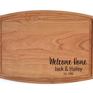 K&ouml;nnte beinhalten: Ein Schneidebrett aus Holz mit abgerundeter Kante und einer personalisierten Nachricht mit dem Text "Willkommen Zuhause Jack & Hailey Est. 2018".