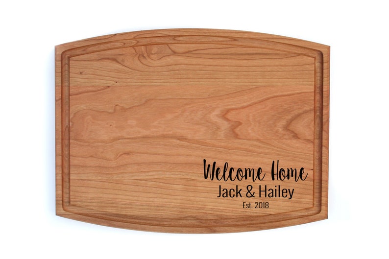 K&ouml;nnte beinhalten: Ein Schneidebrett aus Holz mit abgerundeter Kante und einer personalisierten Nachricht mit dem Text "Willkommen Zuhause Jack & Hailey Est. 2018".