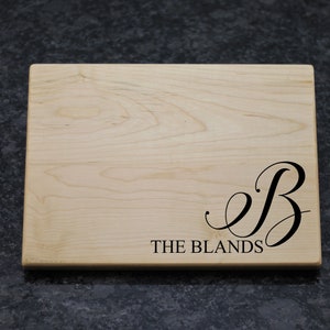 May include: A natural wood cutting board with a black engraved initial "B" and the text "THE BLANDS".