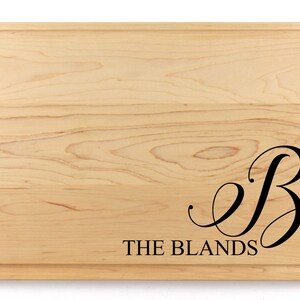 May include: A natural wood cutting board with a rounded edge. The board is engraved with the letter "B" in a cursive font and the text "THE BLANDS".