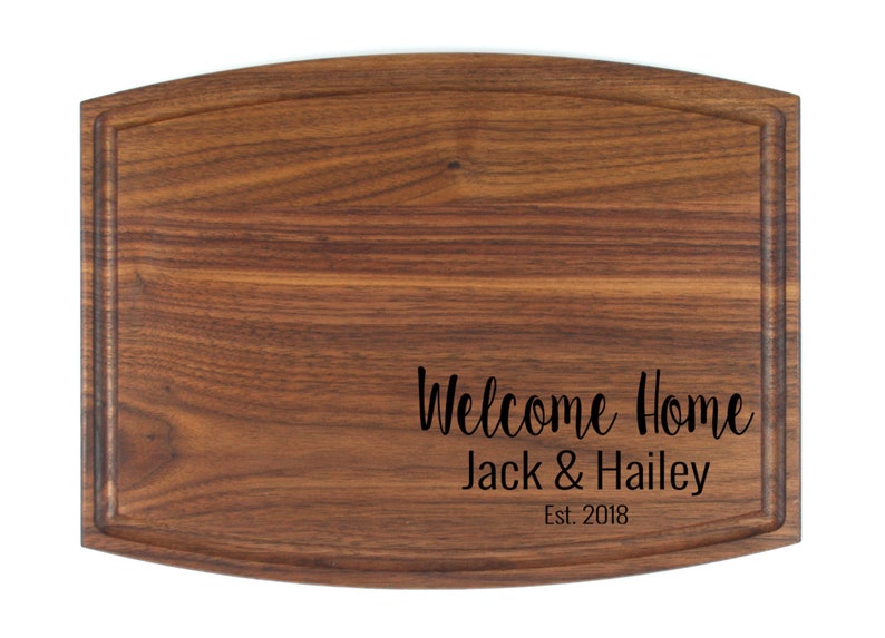 K&ouml;nnte beinhalten: Ein braunes, rechteckiges Schneidebrett aus Holz mit abgerundeten Kanten und einer Rille am Rand. Das Brett ist mit dem Text "Welcome Home Jack & Hailey Est. 2018" graviert.