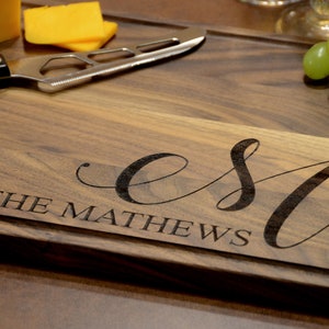 May include: A wooden cutting board with a personalized inscription that reads "EST. THE MATHEWS". The board is brown and has a natural wood grain. There are some cheese wedges and grapes on the board.