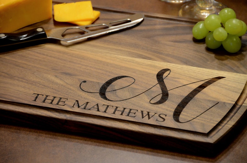 May include: A wooden cutting board with a personalized inscription that reads "EST. THE MATHEWS". The board is brown and has a natural wood grain. There are some cheese wedges and grapes on the board.
