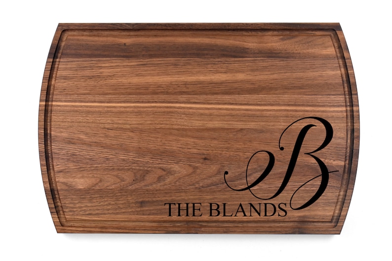 May include: A large, rectangular walnut wood cutting board with a rounded edge. The board is engraved with the letter "B" in a cursive font and the text "THE BLANDS".