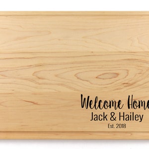 K&ouml;nnte beinhalten: Ein Schneidebrett aus Naturholz mit abgerundeten Kanten und einer personalisierten Inschrift mit dem Text "Willkommen Zuhause Jack & Hailey Est. 2018".