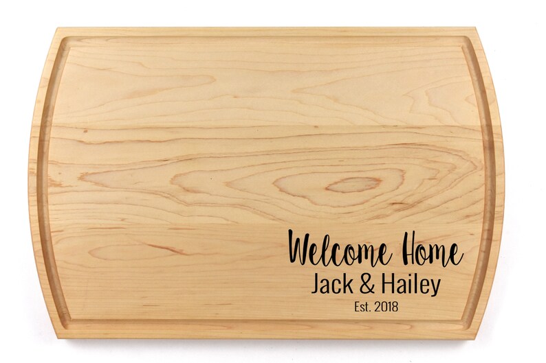 K&ouml;nnte beinhalten: Ein Schneidebrett aus Naturholz mit abgerundeten Kanten und einer personalisierten Inschrift mit dem Text "Willkommen Zuhause Jack & Hailey Est. 2018".