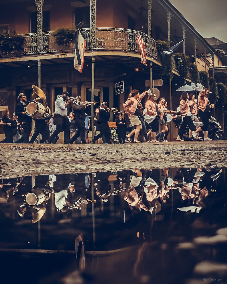 May include: A street scene with a brass band marching in a parade. The band members are wearing dark uniforms and playing instruments. A group of people are walking alongside them. The scene is reflected in a puddle on the ground.