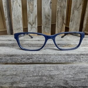 May include: A pair of navy blue eyeglasses with a rectangular frame rests on a weathered wooden surface. The glasses have a classic design with a dark blue frame and clear lenses. The background features a wooden slatted structure.